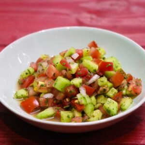 A vibrant cucumber and tomato salad in a white bowl, set on a red wooden table.