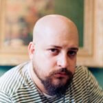 Close-up portrait of a bald man with a beard in a striped shirt, indoors.